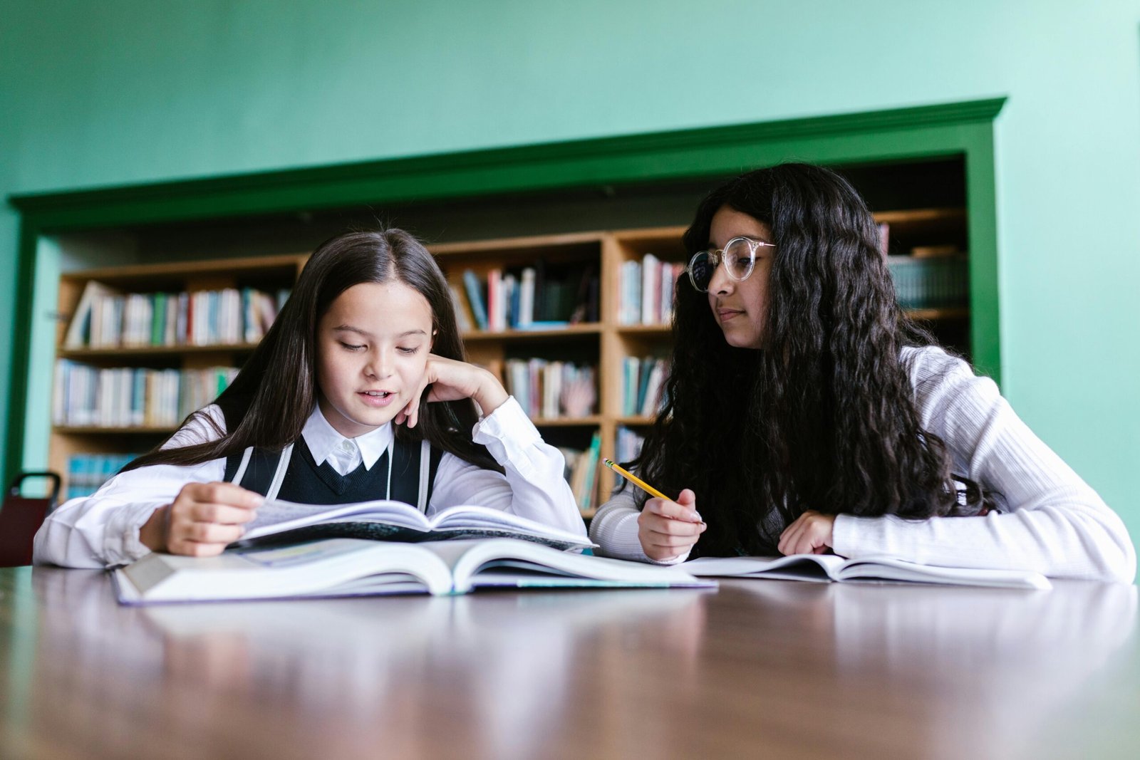 Two girls in school uniforms studying together at a library desk, focused and engaged.